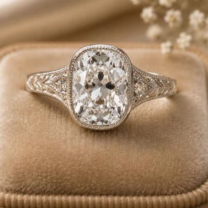 May include: A silver-toned ring featuring a large, cushion-cut center stone set within a milgrain-edged bezel. The band displays intricate, engraved leaf-like patterns along the shoulders, accented by small, embedded round stones on each side.