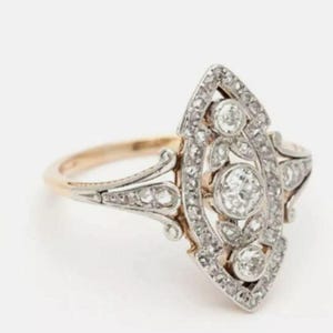 May include: A marquise-shaped ring featuring a central round diamond surrounded by smaller diamonds set in a silver-toned metal frame. The ring has a gold-toned band that tapers into intricate metalwork supporting the elongated diamond-encrusted head.