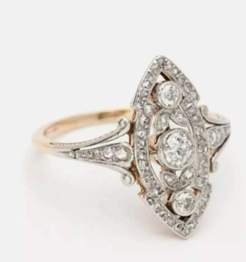 May include: A marquise-shaped ring featuring a central round diamond surrounded by smaller diamonds set in a silver-toned metal frame. The ring has a gold-toned band that tapers into intricate metalwork supporting the elongated diamond-encrusted head.