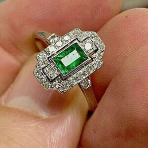 May include: A silver-toned ring featuring a rectangular emerald-cut green gemstone at the center, surrounded by a geometric halo of small, clear, round-cut stones set in a metallic frame with a polished band.