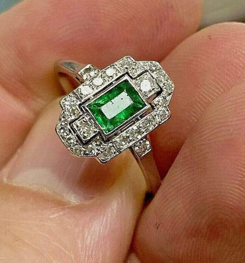 May include: A silver-toned ring featuring a rectangular emerald-cut green gemstone at the center, surrounded by a geometric halo of small, clear, round-cut stones set in a metallic frame with a polished band.