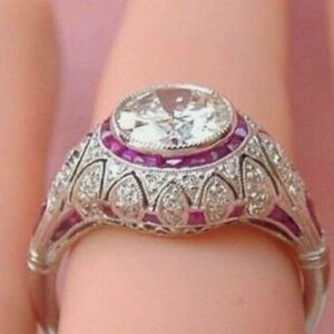 Peut inclure: Une bague argent&eacute;e orn&eacute;e d'une grande pierre centrale ronde et transparente, entour&eacute;e d'un halo de petites pierres rouges &eacute;clatantes. L'anneau et le sertissage pr&eacute;sentent des d&eacute;tails m&eacute;talliques complexes et des pierres transparentes.