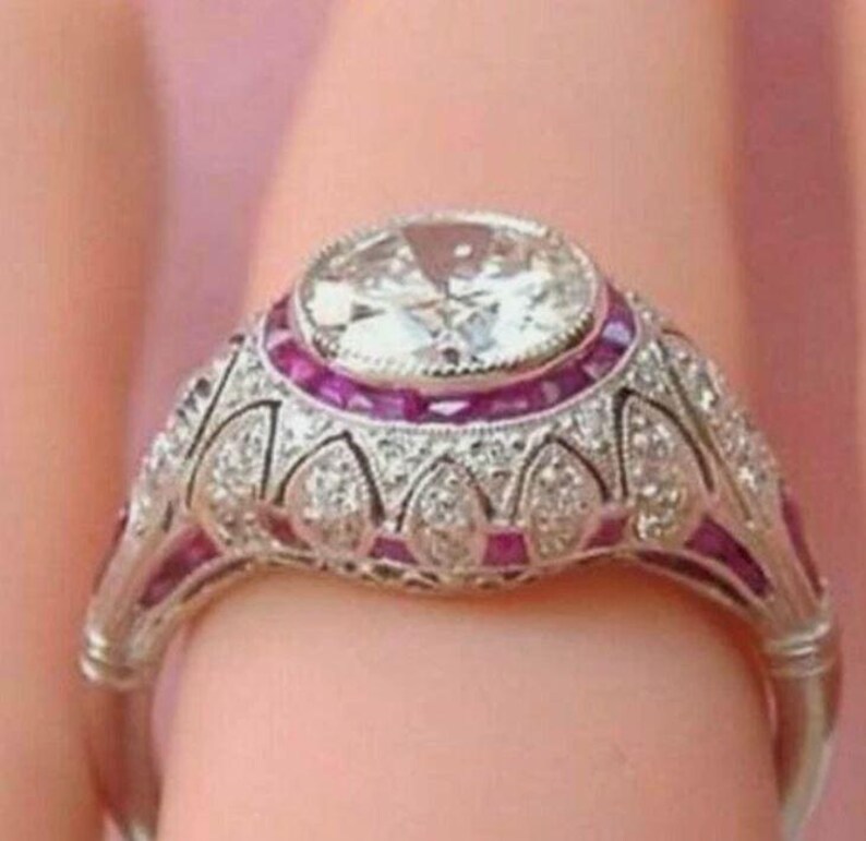 Peut inclure: Une bague argent&eacute;e orn&eacute;e d'une grande pierre centrale ronde et transparente, entour&eacute;e d'un halo de petites pierres rouges &eacute;clatantes. L'anneau et le sertissage pr&eacute;sentent des d&eacute;tails m&eacute;talliques complexes et des pierres transparentes.