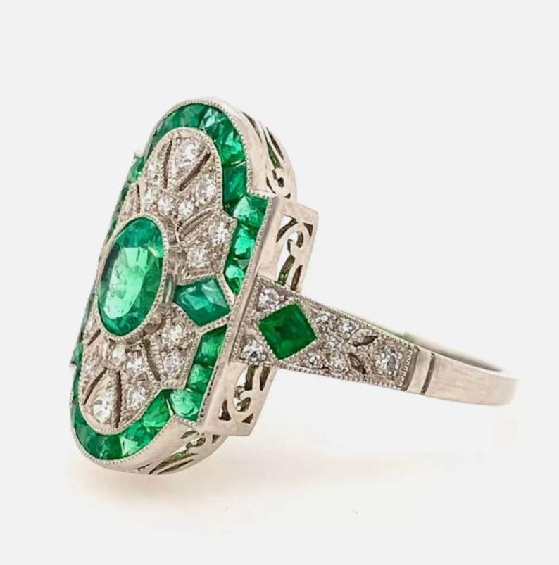 May include: An elongated oval ring crafted from silver-toned metal, featuring a central oval green gemstone surrounded by smaller round white stones and square-cut green stones. The band has intricate metalwork and side-set green accents.