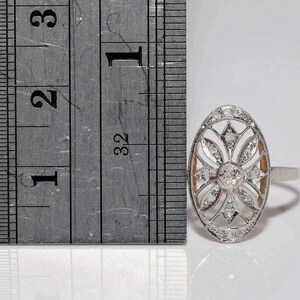 Peut inclure: Une bague ovale de couleur argent&eacute;e pr&eacute;sentant un motif floral ajour&eacute; complexe serti de plusieurs petites pierres claires. Une r&egrave;gle m&eacute;tallique est plac&eacute;e &agrave; c&ocirc;t&eacute; de la bague, montrant que la face ovale mesure environ 1,9 cm de hauteur.