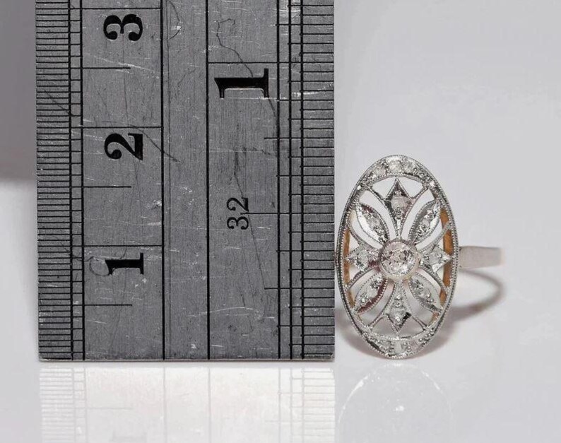 Peut inclure: Une bague ovale de couleur argent&eacute;e pr&eacute;sentant un motif floral ajour&eacute; complexe serti de plusieurs petites pierres claires. Une r&egrave;gle m&eacute;tallique est plac&eacute;e &agrave; c&ocirc;t&eacute; de la bague, montrant que la face ovale mesure environ 1,9 cm de hauteur.