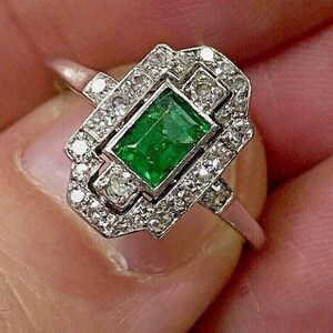 May include: A silver-toned ring featuring a rectangular emerald-cut green gemstone at the center, surrounded by a geometric border of small, clear, round-cut stones set in a metallic frame that extends along the band on both sides.