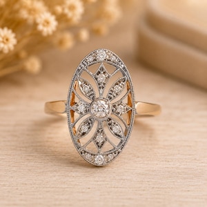 Peut inclure: Une bague de forme ovale avec un diamant rond central entour&eacute; d'une monture en m&eacute;tal filigran&eacute; sertie de plus petits diamants ronds. La bague poss&egrave;de un anneau en or poli reli&eacute; &agrave; la face d&eacute;corative ajour&eacute;e aux tons argent&eacute;s.