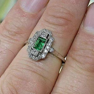 May include: A silver-toned ring featuring a rectangular emerald-cut green gemstone at the center, surrounded by a rectangular halo of small, clear, round-cut stones set in a metallic frame with a thin, polished band.