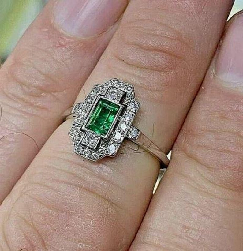 May include: A silver-toned ring featuring a rectangular emerald-cut green gemstone at the center, surrounded by a rectangular halo of small, clear, round-cut stones set in a metallic frame with a thin, polished band.