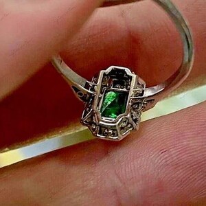 May include: A silver-toned ring featuring a rectangular emerald-green center stone set within a decorative metal frame. The setting is accented with small, clear stones along the sides of the rectangular mount, creating a detailed, geometric design.