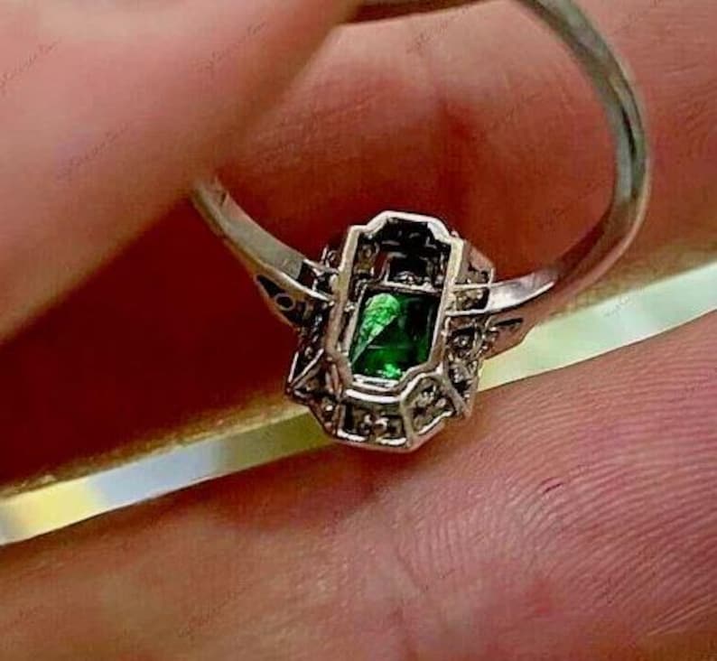 May include: A silver-toned ring featuring a rectangular emerald-green center stone set within a decorative metal frame. The setting is accented with small, clear stones along the sides of the rectangular mount, creating a detailed, geometric design.