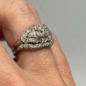 May include: A silver-toned metal ring set consisting of two bands. The primary band features a square-shaped setting with a central round stone surrounded by smaller accent stones. The second band is a curved eternity-style ring with small stones.