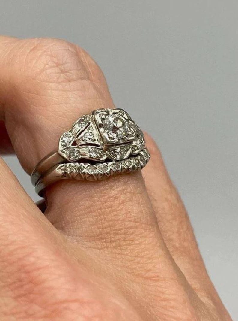 May include: A silver-toned metal ring set consisting of two bands. The primary band features a square-shaped setting with a central round stone surrounded by smaller accent stones. The second band is a curved eternity-style ring with small stones.