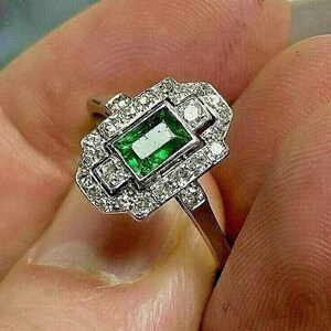 May include: A silver-toned ring featuring a rectangular emerald-cut green gemstone at the center, surrounded by a rectangular halo of small, clear, round-cut stones set in a metallic frame with a polished band.