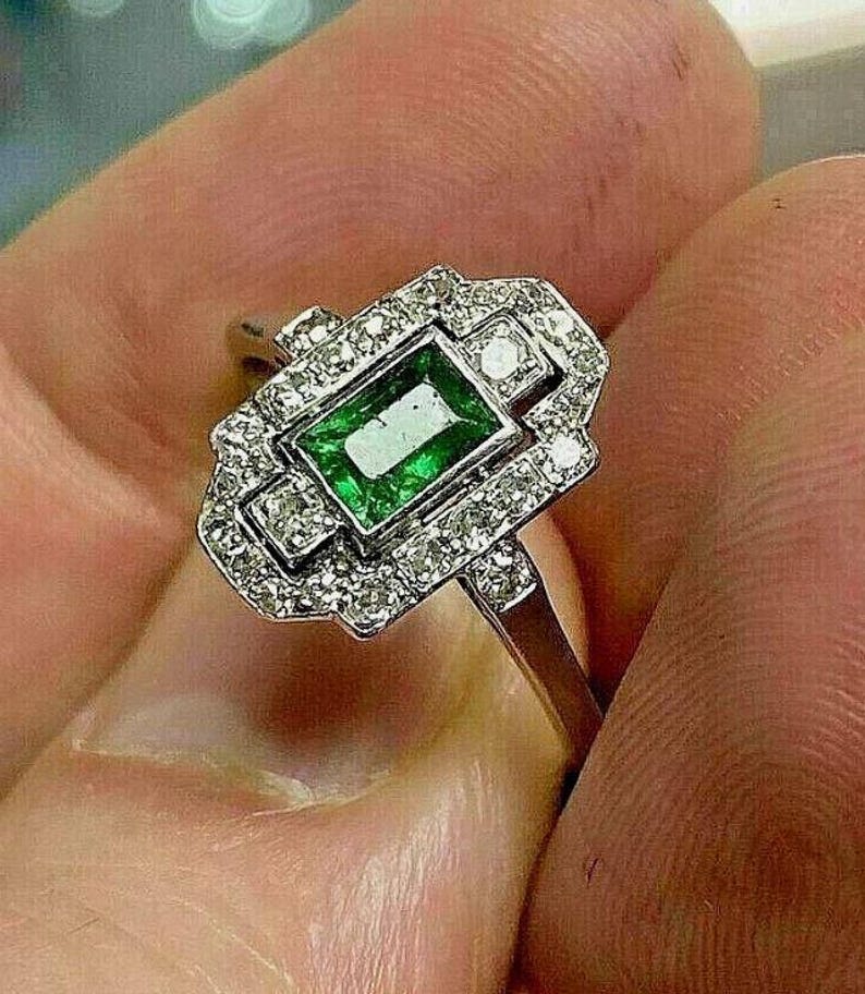 May include: A silver-toned ring featuring a rectangular emerald-cut green gemstone at the center, surrounded by a rectangular halo of small, clear, round-cut stones set in a metallic frame with a polished band.