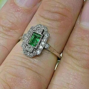 May include: A silver-toned ring featuring a rectangular emerald-cut green gemstone at the center, surrounded by a geometric halo of small, clear round-cut stones. The metal band is slender and polished, supporting the ornate, multi-faceted setting.