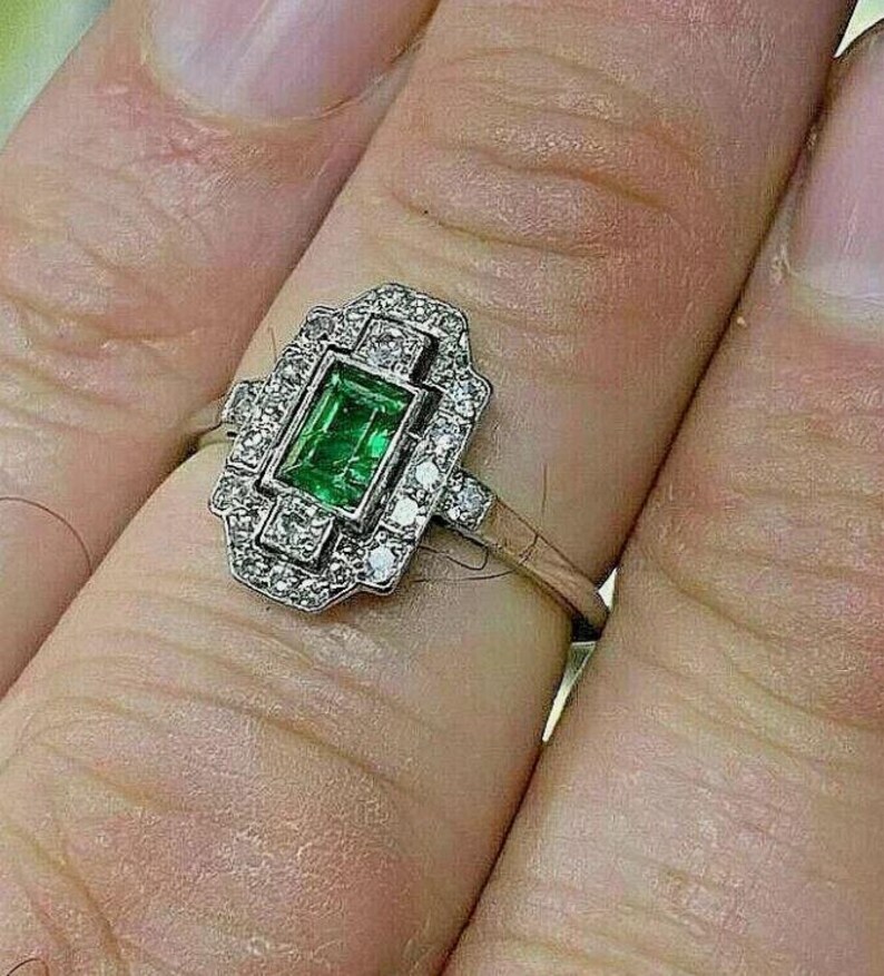 May include: A silver-toned ring featuring a rectangular emerald-cut green gemstone at the center, surrounded by a geometric halo of small, clear round-cut stones. The metal band is slender and polished, supporting the ornate, multi-faceted setting.