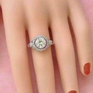 Peut inclure: Une bague argent&eacute;e orn&eacute;e d'une grande pierre centrale ronde et transparente sertie clos, entour&eacute;e d'un halo d&eacute;coratif de petites pierres rondes et transparentes. L'anneau pr&eacute;sente des d&eacute;tails m&eacute;talliques complexes et d'autres petites pierres.