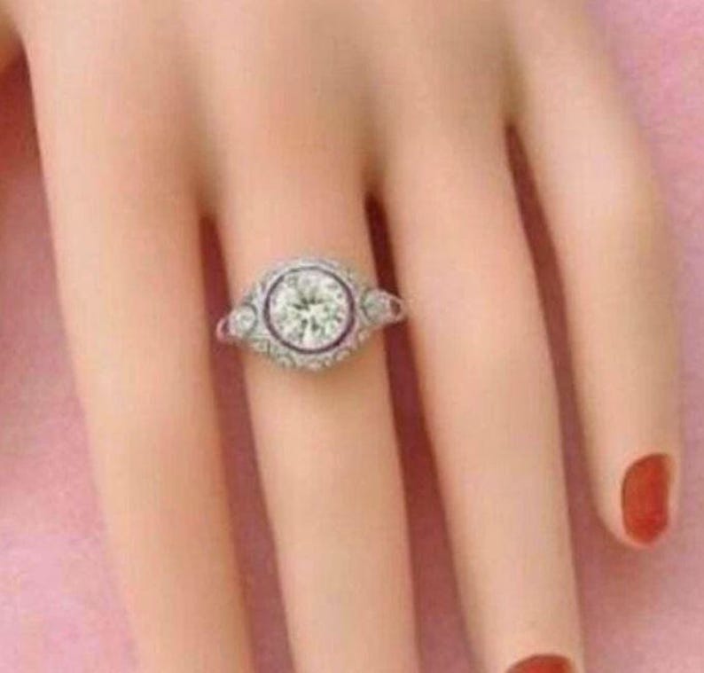 Peut inclure: Une bague argent&eacute;e orn&eacute;e d'une grande pierre centrale ronde et transparente sertie clos, entour&eacute;e d'un halo d&eacute;coratif de petites pierres rondes et transparentes. L'anneau pr&eacute;sente des d&eacute;tails m&eacute;talliques complexes et d'autres petites pierres.
