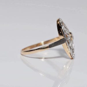 Peut inclure: Une bague dot&eacute;e d'un anneau en or qui se transforme en une monture en m&eacute;tal sombre. La face de la bague est une plaque allong&eacute;e en forme de losange, incrust&eacute;e de multiples petites pierres claires et scintillantes dispos&eacute;es en motif sym&eacute;trique.