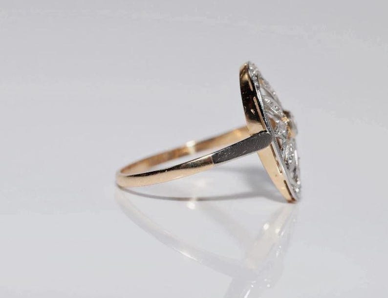 Peut inclure: Une bague dot&eacute;e d'un anneau en or qui se transforme en une monture en m&eacute;tal sombre. La face de la bague est une plaque allong&eacute;e en forme de losange, incrust&eacute;e de multiples petites pierres claires et scintillantes dispos&eacute;es en motif sym&eacute;trique.