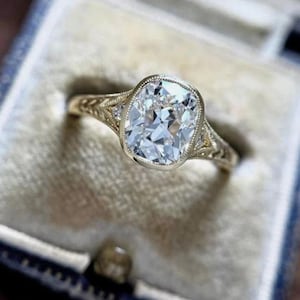 May include: A gold ring featuring a large, cushion-cut center stone set in a bezel with milgrain detailing. The band displays intricate, openwork metal patterns along the shoulders, accented by small, round, clear stones on each side.