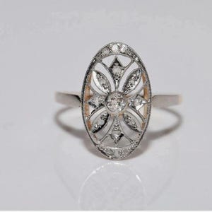 Peut inclure: Une bague de forme ovale au ton argent&eacute; pr&eacute;sentant un design complexe en filigrane ajour&eacute;. Le centre est orn&eacute; d'une pierre ronde transparente, entour&eacute;e de petites pierres transparentes dispos&eacute;es en motif floral sur la monture allong&eacute;e.