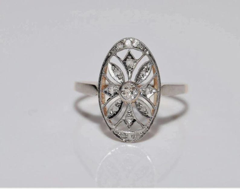 Peut inclure: Une bague de forme ovale au ton argent&eacute; pr&eacute;sentant un design complexe en filigrane ajour&eacute;. Le centre est orn&eacute; d'une pierre ronde transparente, entour&eacute;e de petites pierres transparentes dispos&eacute;es en motif floral sur la monture allong&eacute;e.