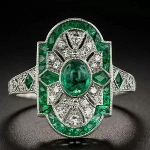 May include: A silver-toned ring featuring a central oval-cut green gemstone surrounded by a geometric arrangement of smaller green stones and clear white stones. The metal band is detailed with intricate patterns and a symmetrical design.