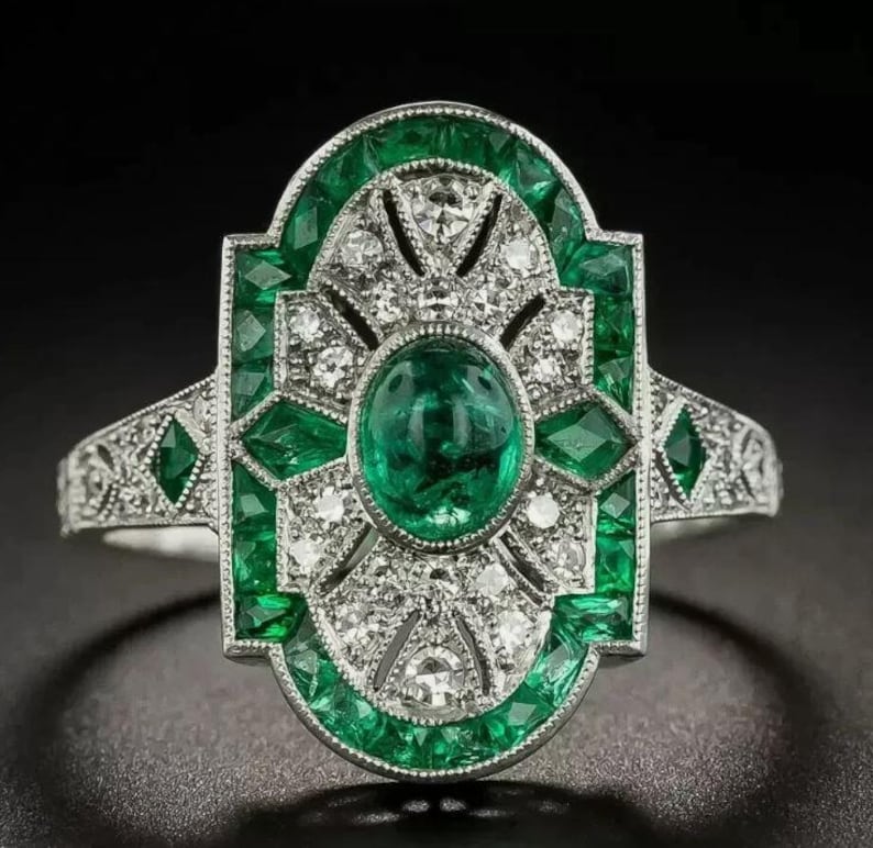 May include: A silver-toned ring featuring a central oval-cut green gemstone surrounded by a geometric arrangement of smaller green stones and clear white stones. The metal band is detailed with intricate patterns and a symmetrical design.