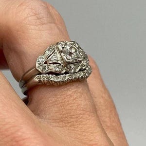 May include: A silver-toned metal ring set consisting of two bands. The primary band features a square-shaped setting with a central round stone surrounded by smaller accent stones. The second band is a curved eternity-style ring with small stones.