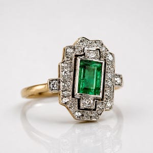 May include: A gold ring featuring a central rectangular emerald gemstone surrounded by a geometric halo of small round diamonds. The setting is crafted with a silver-toned metal frame around the stones, contrasting with the gold band.