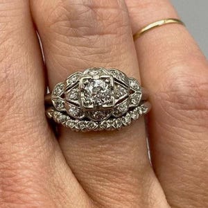 May include: A silver-toned engagement ring set featuring a central square-set diamond surrounded by a decorative filigree frame with smaller round diamonds, paired with a curved wedding band encrusted with a row of small round diamonds.