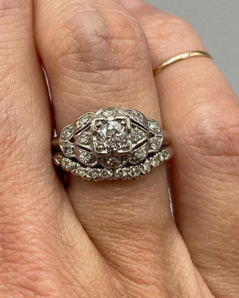 May include: A silver-toned engagement ring set featuring a central square-set diamond surrounded by a decorative filigree frame with smaller round diamonds, paired with a curved wedding band encrusted with a row of small round diamonds.