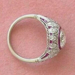 Peut inclure: Une bague argent&eacute;e orn&eacute;e d'une pierre centrale ronde entour&eacute;e d'un halo de petits diamants et de pierres rouges &eacute;clatantes. L'anneau pr&eacute;sente un travail de m&eacute;tal en filigrane complexe avec un motif r&eacute;p&eacute;titif d'accents en diamant.