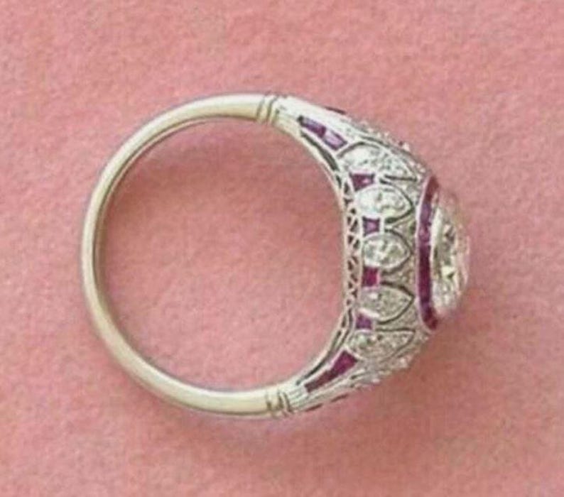Peut inclure: Une bague argent&eacute;e orn&eacute;e d'une pierre centrale ronde entour&eacute;e d'un halo de petits diamants et de pierres rouges &eacute;clatantes. L'anneau pr&eacute;sente un travail de m&eacute;tal en filigrane complexe avec un motif r&eacute;p&eacute;titif d'accents en diamant.
