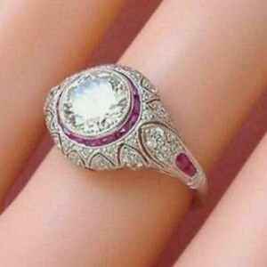 Peut inclure: Une bague argent&eacute;e orn&eacute;e d'une grande pierre centrale ronde et transparente, entour&eacute;e d'un halo de petites pierres rouges &eacute;clatantes. L'anneau en m&eacute;tal est finement d&eacute;taill&eacute; avec de petites pierres blanches scintillantes en filigrane.