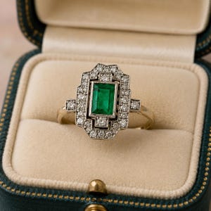 May include: A gold ring featuring a central emerald-cut green gemstone surrounded by a rectangular halo of small round white diamonds. The setting is geometric with a stepped design, and the band is a thin, polished gold metal.