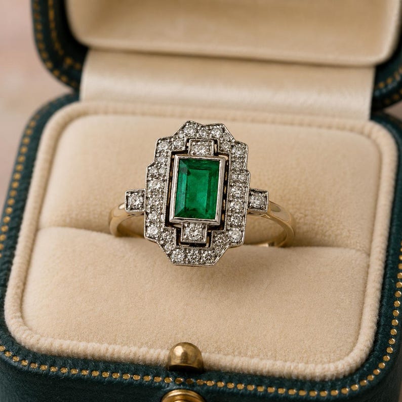 May include: A gold ring featuring a central emerald-cut green gemstone surrounded by a rectangular halo of small round white diamonds. The setting is geometric with a stepped design, and the band is a thin, polished gold metal.