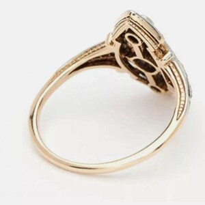 May include: A gold ring featuring an elongated, marquise-shaped head with a central openwork design. The band is smooth and polished, while the setting displays intricate metalwork and small, clear stones along the outer edges of the frame.