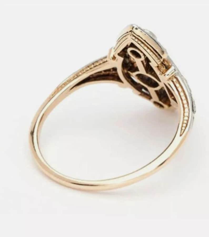 May include: A gold ring featuring an elongated, marquise-shaped head with a central openwork design. The band is smooth and polished, while the setting displays intricate metalwork and small, clear stones along the outer edges of the frame.