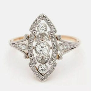 May include: A marquise-shaped ring featuring a central round diamond flanked by two smaller round diamonds, all set in a silver-toned metal frame. The elongated setting is accented with small pav&eacute;-set stones and leaf-like metal details.