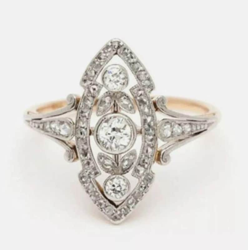 May include: A marquise-shaped ring featuring a central round diamond flanked by two smaller round diamonds, all set in a silver-toned metal frame. The elongated setting is accented with small pav&eacute;-set stones and leaf-like metal details.