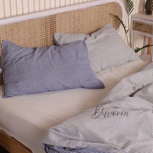May include: A bedding set featuring a white fitted sheet and a duvet cover with a blue and white pinstripe pattern. Two matching rectangular pillows with the same pinstripe design are placed against a headboard with a woven cane insert. The text Duvoria is printed on the duvet cover.