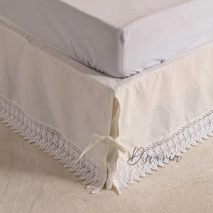 May include: A cream-colored bed skirt featuring a delicate white lace trim along the bottom edge and side ties for adjustment. The fabric is smooth and opaque, draped over the base of a bed with a white mattress cover visible on top.