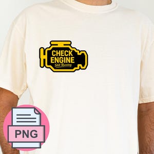May include: A cream-colored short-sleeve t-shirt featuring a graphic on the left chest. The graphic is a yellow and black engine icon with the text "CHECK ENGINE" and "Still Running" printed in black inside the engine shape.