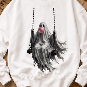 May include: A white crewneck sweatshirt featuring a central graphic of a ghost draped in a white sheet, sitting on a swing suspended by black chains. The ghost has large dark eye sockets and is blowing a round, bright pink bubble with gum.