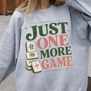 Könnte beinhalten: Ein graumeliertes Sweatshirt mit Rundhalsausschnitt und einem Grafikdruck mit der Aufschrift JUST ONE MORE GAME in dunkelgrüner und gedeckter roter Schrift. Das Design zeigt einen Stapel aus vier Mahjong-Steinen mit Symbolen und einer Blume.