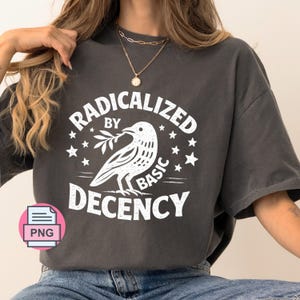 以下が含まれることがあります： チャコールグレーの半袖Tシャツ。枝をくわえた鳥と星が描かれた白いグラフィックが中央にあり、RADICALIZED BY BASIC DECENCYという文字がかすれた白のフォントでプリントされています。ゴールドのネックレスを合わせて着用しています。